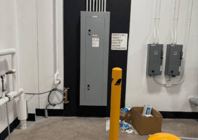 A gray electrical panel mounted on a black wall, with conduit pipes above, yellow safety bollards in front, and electrical supplies in an open cardboard box nearby.