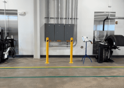 Industrial workspace with exposed conduit pipes, electrical panels on the wall, yellow safety bollards, machinery, and a stand with a tablet.