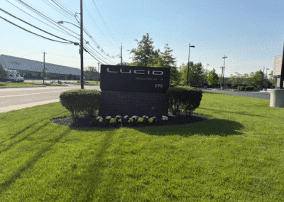 A black sign reading "LUCID SERVICE DROP OFF" stands on a lawn with trimmed bushes, flowers, and a road in the background.