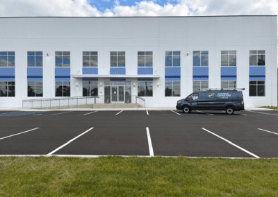 A white commercial building with blue accents, empty parking spaces, and a dark van parked near the entrance under a partly cloudy sky.