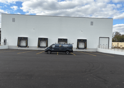 A black van is parked in front of a white industrial building with five loading docks on a newly paved lot under a partly cloudy sky.